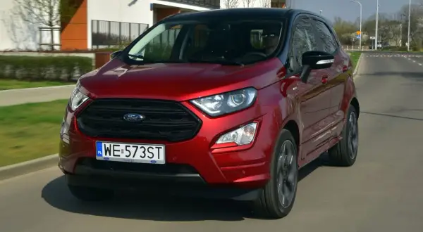 Czerwony Ford Ecosport w ruchu miejskim. Kierowca w okularach przeciwsłonecznych. Wiele pozytywnych ford ecosport opinie potwierdza jego miejski charakter.