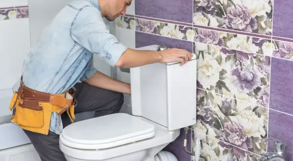 Instalator w pasie z narzędziami montuje biały stelaż podtynkowy do toalety. Łazienka ozdobiona jest fioletowymi płytkami z kwiatowym wzorem.