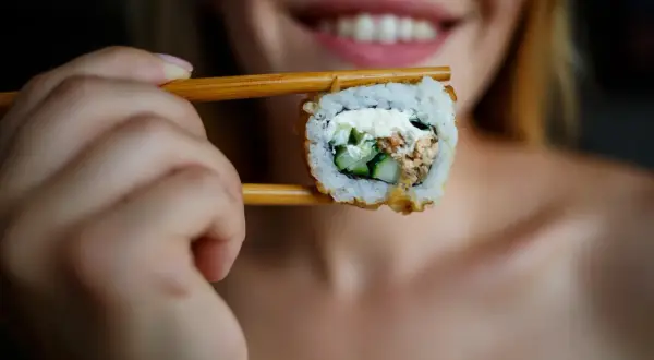 Uśmiechnięta kobieta trzyma pałeczkami sushi, zastanawiając się, czy można jeść sushi karmiąc piersią.