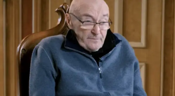 Phil Collins w okularach, siedzi w fotelu. Jego choroba wpływa na jego zdolność do gry na perkusji, ale nadal inspiruje fanów.
