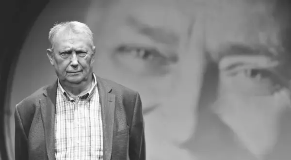Wojciech Młynarski, jakby z tekstu "Jesteśmy na wczasach", w marynarce i kratowanej koszuli, na tle swojego portretu.