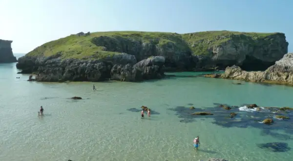 Gente disfrutando del agua cristalina en la costa de Llanes, con acantilados verdes y rocosos de fondo.