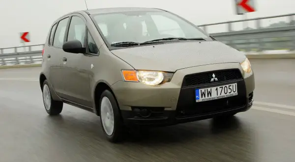 Złoty Mitsubishi Colt 2006 na mokrej drodze, światła włączone, w tle znaki drogowe.
