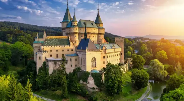 Bajkowy zamek otoczony zielenią, z wieżami i fosą. Słońce zachodzi, malując niebo na różowo i pomarańczowo. To idealne miejsce na magiczną noc w zamku.