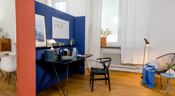 Ein blauer Raumteiler trennt den Arbeitsplatz im Wohnzimmer ab. Ein Schreibtisch mit Laptop und Lampe steht bereit.
