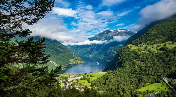 Widok na fiord podczas rejsu do Norwegii. Statek wycieczkowy płynie po spokojnej wodzie, otoczonej zielonymi górami i chmurami.