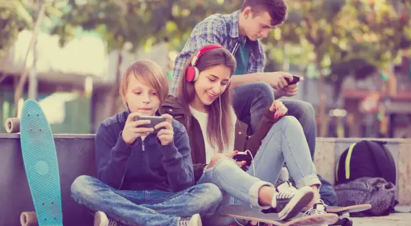 Trójka nastolatków pochłonięta smartfonami, obok deskorolki. Czy to początek uzależnień behawioralnych?