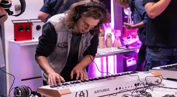 Młody mężczyzna w słuchawkach gra na keyboardzie Arturia. W tle widać sprzęt audio i innych ludzi, tworząc atmosferę pracy w wytwórni muzycznej.