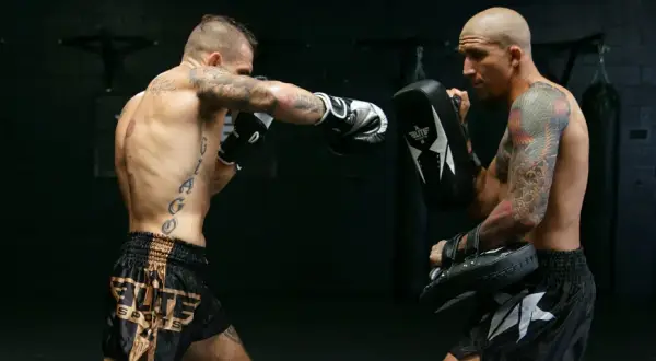 Two men practice boxing without sparring. One throws a punch while the other holds a pad to catch it.