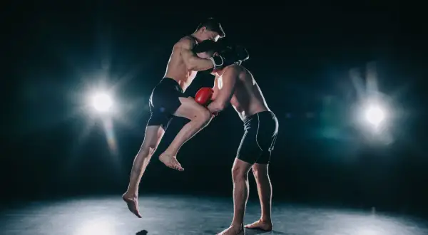Two boxers spar intensely. One leaps, delivering a knee strike while the other defends. This dynamic action shows how boxers do calisthenics for explosive power.