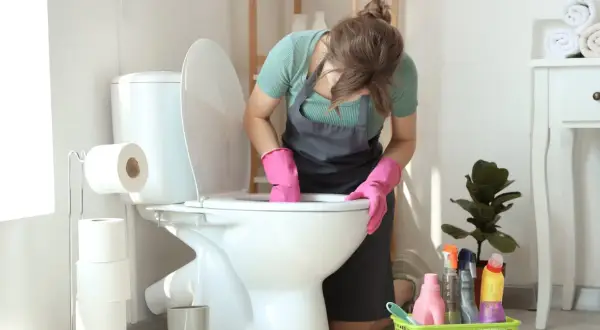 Kobieta w różowych rękawiczkach czyści toaletę, bo w łazience smierdzi. Obok stoją środki czystości i rolki papieru toaletowego.