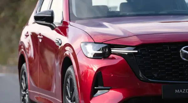 Błyszczący, czerwony lakier nowej Mazdy CX-60 odbija niebo. Widoczny fragment maski, reflektor i charakterystyczny grill.