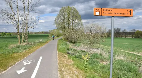 Rowerzysta jedzie ścieżką rowerową przez zielone pola. Pomarańczowy znak wskazuje kierunek do Kotbacza i Szczecin-Śmirdnica.
