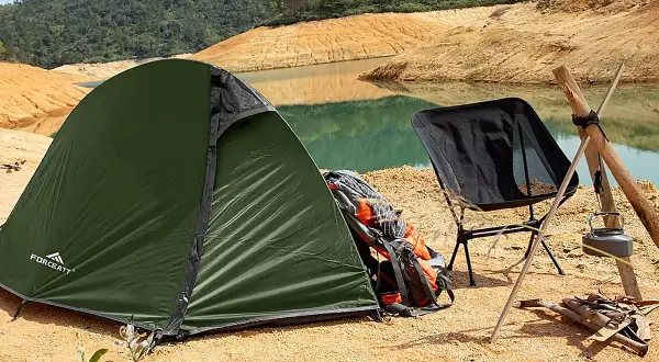 Tienda de campaña verde junto a un lago, con una silla plegable negra y mochilas de transporte.