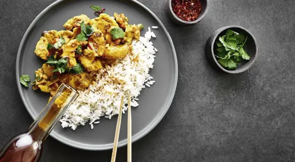 Ein Teller mit Reis und einem köstlichen Thai Curry, garniert mit frischem Koriander. Daneben kleine Schälchen mit Chiliflocken und Koriander.