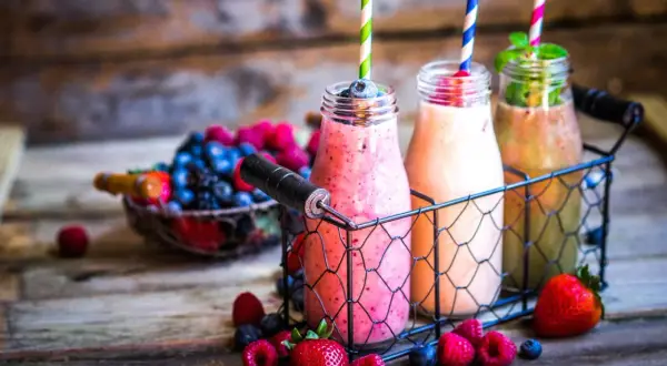 Drei bunte Flaschen mit leckeren Smoothies zum Abnehmen, umgeben von frischen Beeren und Erdbeeren auf rustikalem Holz.