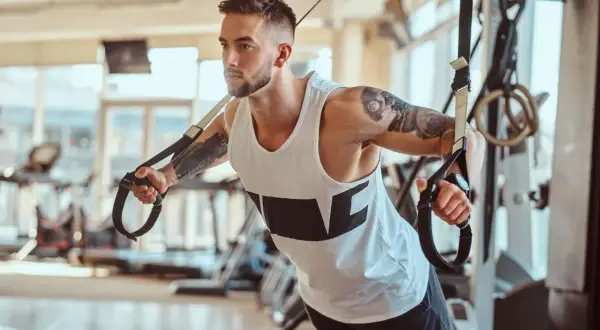 Mężczyzna z tatuażami ćwiczy na taśmach TRX. Zastanawiasz się, ile trwa trening na siłowni? Ten intensywny wysiłek to klucz do sukcesu.
