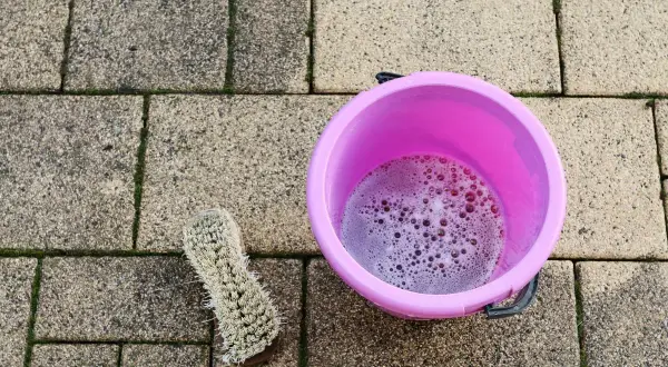 Różowe wiaderko z pianą i szczotka na kostce brukowej. Czas na czyszczenie kostki brukowej octem, by pozbyć się mchu.
