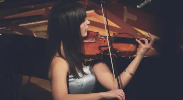 Eine Frau spielt Geige, ihr Bogen erzeugt einen zarten vibrato geige Klang. Im Hintergrund ist ein Klavier zu sehen.