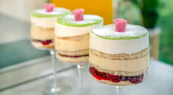 Tre vackra prinsesstårtor i glas, med lager av sockerkaka, grädde, vaniljkräm och hallonsylt, toppade med grön marsipan och en rosa ros.