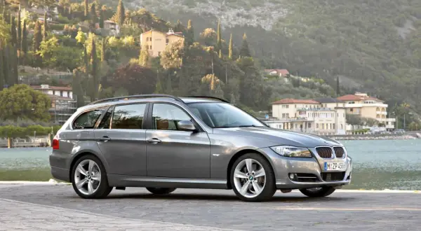 Szary kombi BMW 325i zaparkowany nad jeziorem, z malowniczymi wzgórzami i budynkami w tle.