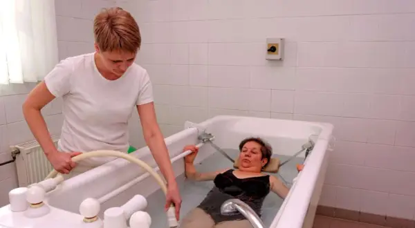 Therapeutin führt Balneotherapie bei Arthrose durch. Eine Frau liegt in einem Wasserbad, während die Therapeutin eine Düse hält.