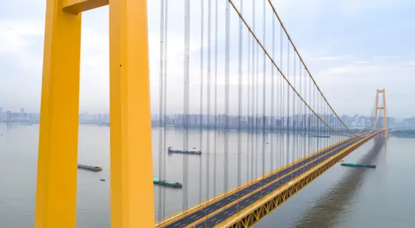 Żółty, wiszący most w Chinach przecina szeroką rzekę, po której płyną barki. W oddali widać panoramę miasta.