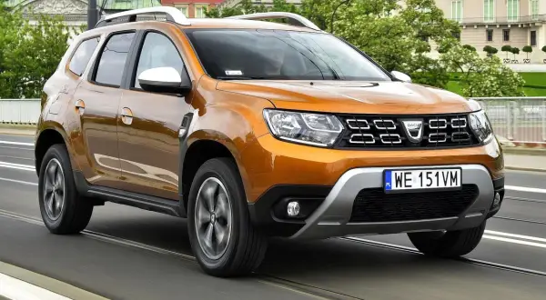 Pomarańczowy Dacia Duster 2 jedzie po moście, za nim widać budynki i drzewa.