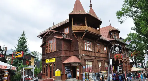 Drewniana architektura na Krupówkach, z charakterystycznymi zdobieniami i wieżyczkami. W tle widać ludzi spacerujących po tętniącej życiem ulicy.