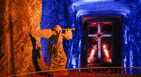 Statua di un angelo con tromba nella cattedrale del sale, illuminata da luci blu e arancioni, con una croce sullo sfondo.
