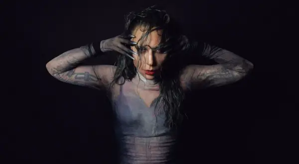 Lady Gaga w siateczkowej bluzce z długimi rękawami i tatuażami, zakrywająca twarz dłońmi.