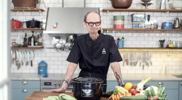 En kock med tatueringar står vid ett köksbord med en slow cooker och färska grönsaker. Perfekt för att hitta nya ångkokare recept.