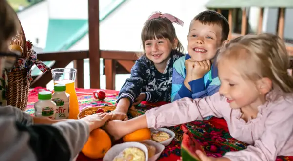 Radosna majówka w górach z dziećmi. Uśmiechnięte maluchy przy stole pełnym owoców, kefirów i słodkości.