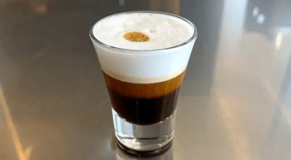 Kawa espresso corretto z pianką i kroplą likieru, podana w szklance na metalowym blacie.