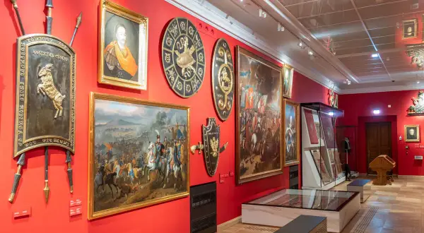 Wnętrze muzeum w Krakowie z obrazami historycznymi, portretem i zbrojami na czerwonych ścianach.