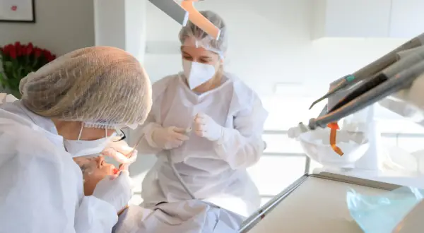 Dentystka w masce i fartuchu wykonuje zabieg kiretażu u pacjentki. W tle widać sprzęt stomatologiczny.