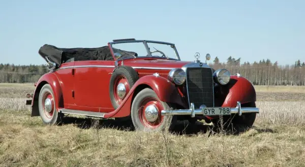Röd cabriolet, en av de äldre tyska bilarna, parkerad på en äng med skog i bakgrunden.