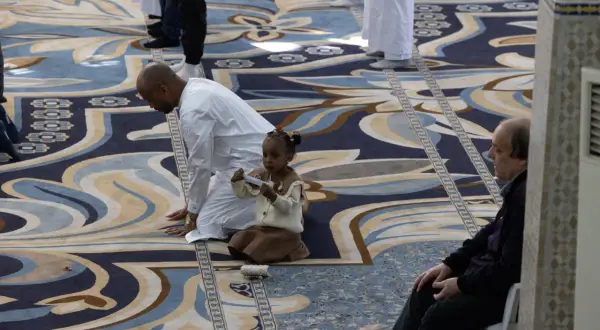 Bambina e uomini in preghiera su un tappeto decorato, un momento di pace durante il Ramadan a Roma.