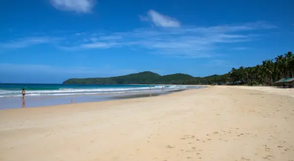 Słoneczny Nacpan Beach z turkusową wodą, zielonymi wzgórzami i palmami. Kilka osób spaceruje po piaszczystym brzegu.