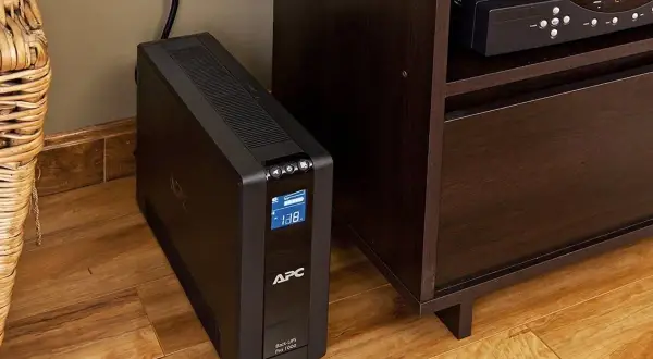 Czarny UPS APC stoi na drewnianej podłodze obok mebla. Jaki UPS, chroni sprzęt przed zanikiem prądu.
