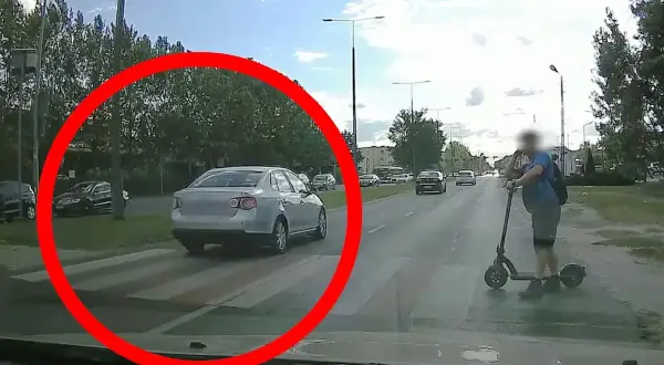 Srebrny samochód na pasach, wymuszenie pierwszeństwa. Na hulajnodze stoi mężczyzna.