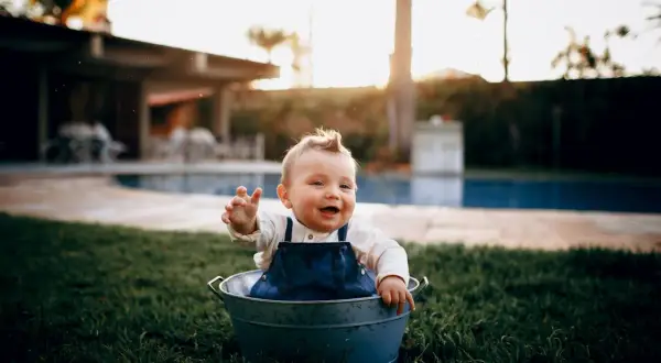Glad bebis i en hink på gräsmattan, med en pool i bakgrunden. En härlig dag för ett bebis bada i poolen!