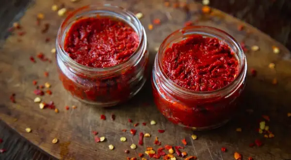 Dwa słoiczki z adżiką, idealną do doprawienia dań. Na drewnianej desce rozsypane płatki chili.