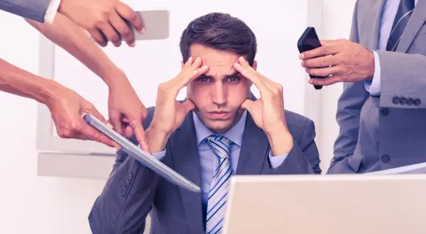 Stressad man med händer på tinningarna, omgiven av kollegor som ger instruktioner. Dålig kognitiv ergonomi leder till överbelastning.