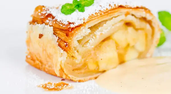 Ein Stück Apfelstrudel mit Puderzucker und Minze, serviert mit Vanillesauce.