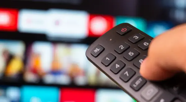 Dłoń trzyma pilot, a na ekranie telewizora widać kolorowe kafelki z filmami. Zastanawiasz się, iptv co to? To przyszłość telewizji!