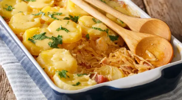 Zapiekanka z ziemniaków i kapusty w naczyniu, ozdobiona natką pietruszki, z drewnianymi łyżkami.