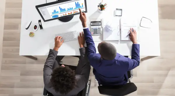 Två personer vid ett skrivbord analyserar data på en skärm, ett exempel på it-system. En person i rullstol skriver på tangentbordet, den andra pekar på diagrammet.