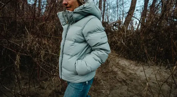 Kobieta w jasnoszarej, puchowej kurtce z kapturem Columbia Puffect Jacket spaceruje po leśnej ścieżce.