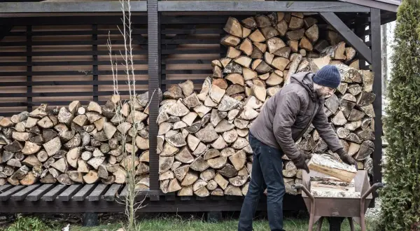 Mann lagert Holz im Freien. Ein Mann mit Bart und Mütze legt Holzscheite in eine Schubkarre, daneben ein großer Stapel Brennholz.
